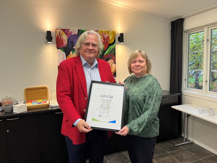 En vithårig man i röd kavaj tar emot ett diplom av en kvinna i grön blus.