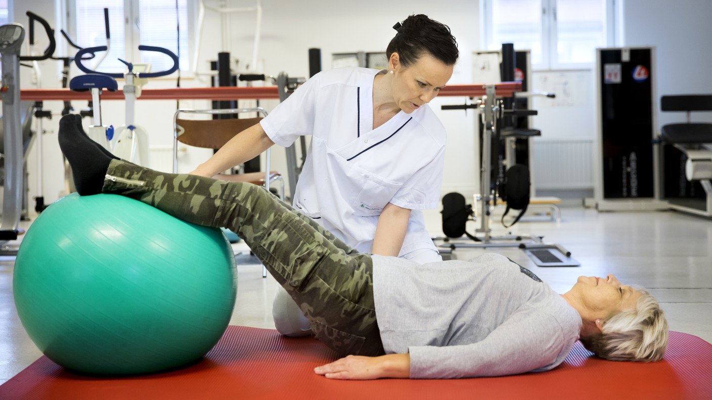 En sjukgymnast hjälper en kvinna  som ligger på rygg och har fötterna på en pilatesboll.