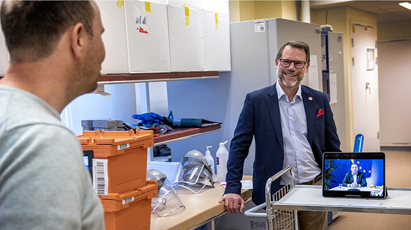 Erik Fredholm och Andreas Svahn står och pratar mitt emot varandra. Stefan Löfven finns med på bilden via en länk på en dator.