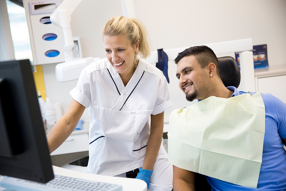 Tandhygienist pratar med en man som sitter i behandlingsstol. De tittar tillsammans på en datorskärm.