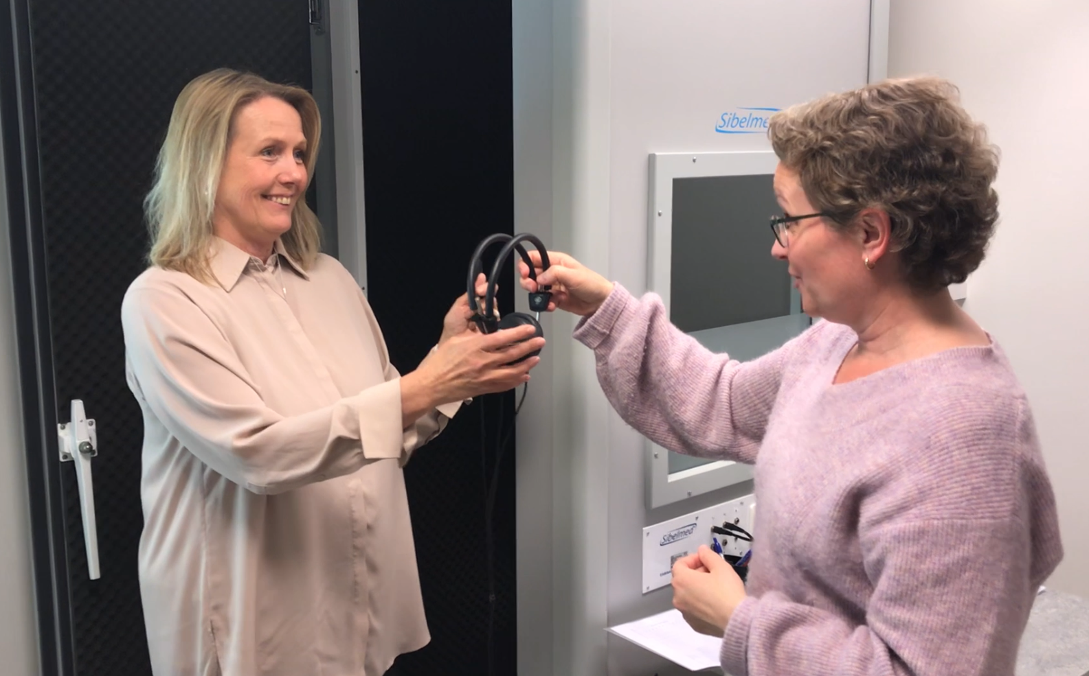 Två kvinnor står mitt emot varandra där den ena räcker den andre ett par hörlurar