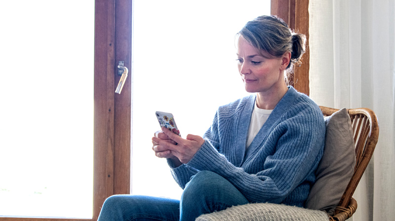 Kvinna sitter uppkrupen i en fåtölj med en mobiltelefon i handen. Hon ler lätt. 