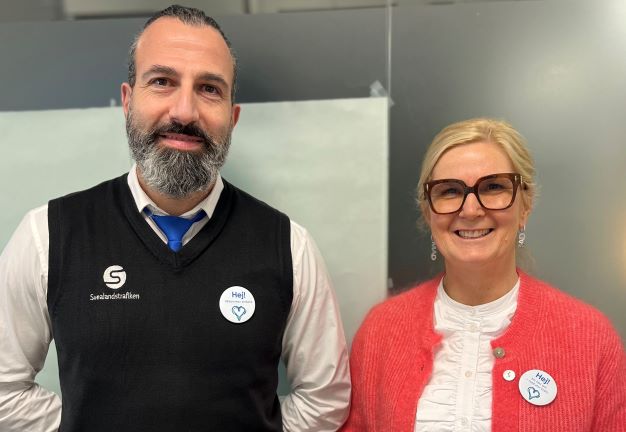 En skäggig man och en kvinna med glasögon. Båda ler och har en knapp med HEJ på sig.